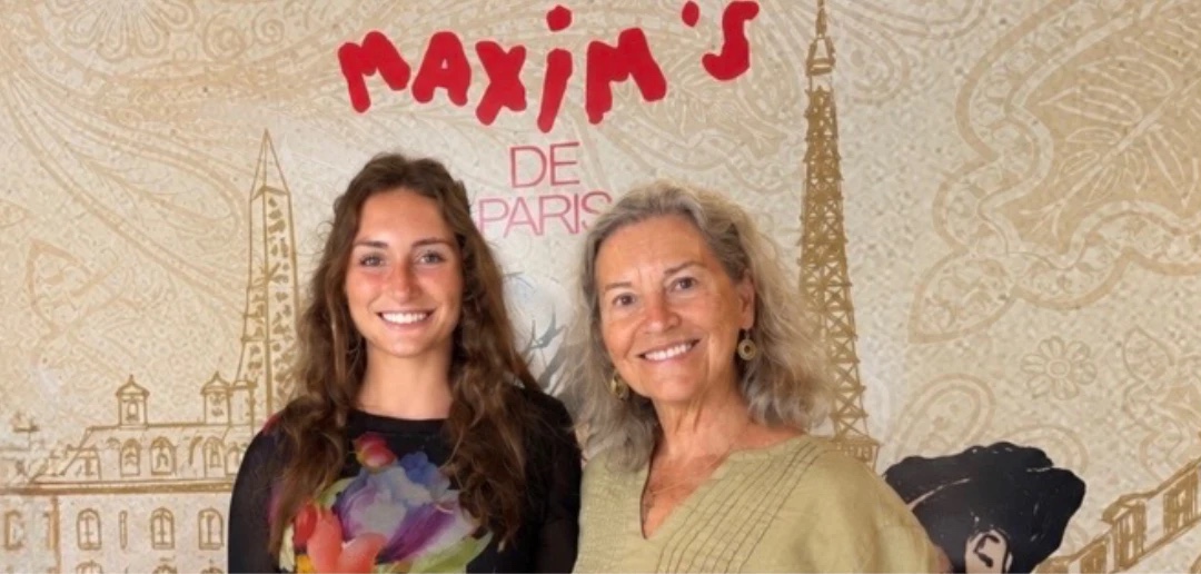 Kate Adams and Terri Birkhauser smiling in front of a Maxim's de Paris mural
